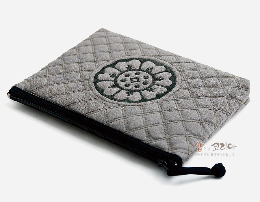 [텍스트: 샵오브코리아 대한민국의 품격까지 전합니다] 회색 누비 원단 위 흑색 백제 수막새 연꽃 문양 자수와 블랙 지퍼 장식이 어우러진 직사각형 파우치 디자인