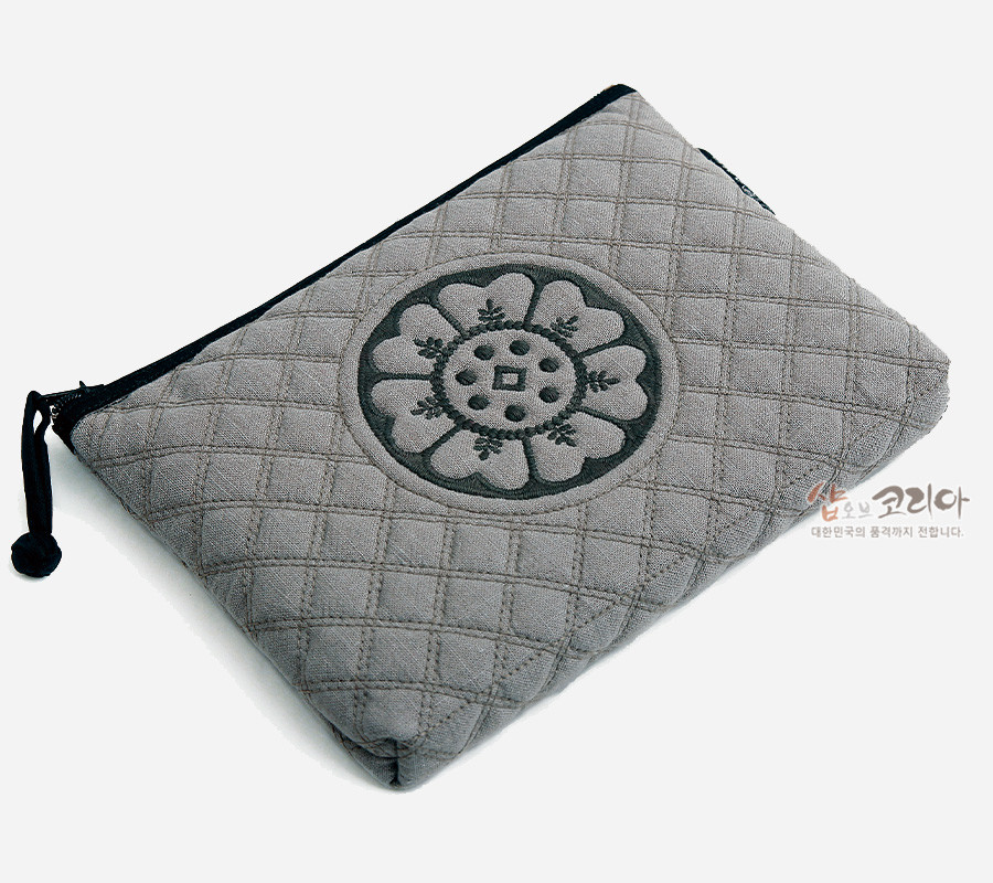 [텍스트: 샵오브코리아 대한민국의 품격까지 전합니다.] 그레이 누비 패브릭 위에 흑색 백제 수막새 연꽃 문양 자수 장식, 블랙 지퍼와 끈이 더해진 직사각형 파우치 디자인