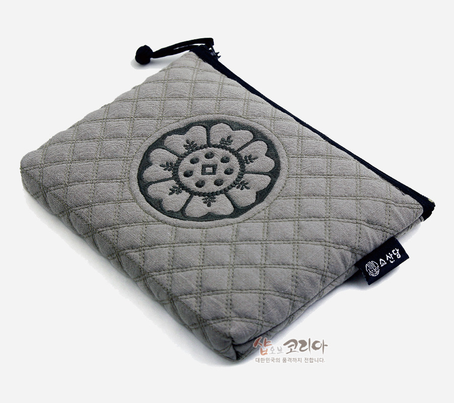 [텍스트: 샵오브코리아 대한민국의 품격까지 전합니다 / 소산당] 회색 누비 원단에 흑색 백제 수막새 연꽃 문양 자수 장식, 흑색 지퍼와 끈으로 마감한 사각 파우치 형태