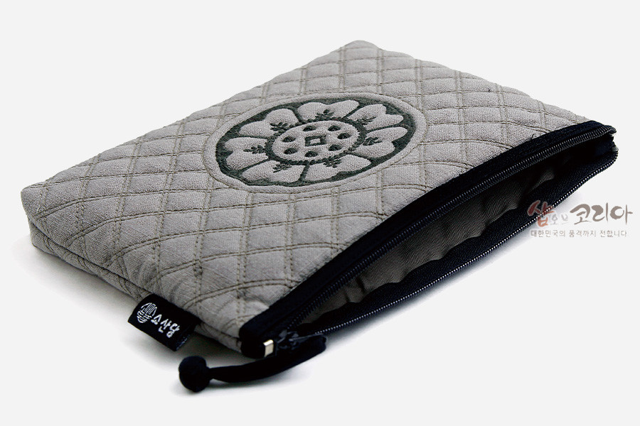 [텍스트: 샾오브코리아] 회색 누비 원단 위 백제 수막새 연꽃 문양 흑색 자수 장식, 블랙 지퍼와 끈 디테일의 직사각 파우치 디자인