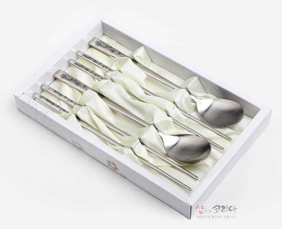 [텍스트: 샵오브코리아 대한민국의 품격까지 전합니다.] 스테인리스 재질, 손잡이에 매화 문양이 섬세하게 양각된 은색 전통 숟가락·젓가락 2인 세트, 아이보리색 패브릭 받침과 화이트 박스 구성 이미지 설명