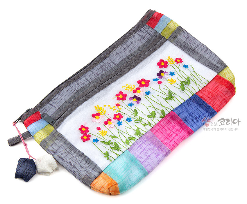 [텍스트: 샵오브코리아 대한민국의 품격까지 전합니다.] 모시 소재에 오방색 조각 천을 누빔하여 두른 테두리와 다채로운 들꽃 야생화 자수, 지퍼 장식과 매듭 달린 패브릭 참 장식 파우치 이미지 설명