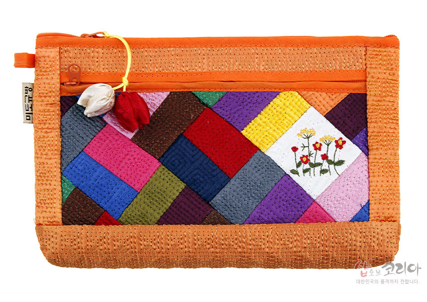 [텍스트: 샵오브코리아 대한민국의 품격까지 전합니다.] 주황색 누비 바탕에 다채로운 비단 조각보 누비 기법과 흰색 조각 위 꽃자수 문양, 조개 모양 매듭 노리개 장식이 어우러진 파우치 이미지 설명