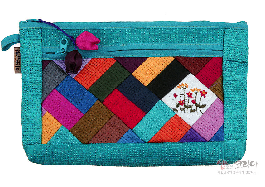 [텍스트: 샵오브코리아 대한민국의 품격까지 전합니다.] 하늘색 누비 원단 위 알록달록 조각보 패치워크와 꽃자수 문양이 조화를 이룬 지퍼형 파우치 이미지 설명