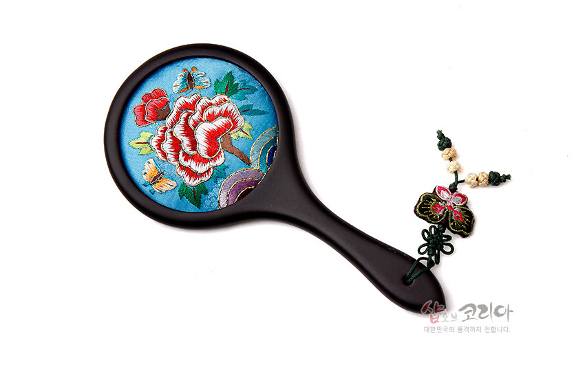 [텍스트: 샵오브코리아 대한민국의 품격까지 전합니다.] 하늘색 비단 바탕에 붉은 꽃과 나비 자수가 놓인 원형 손거울, 어두운 금속 손잡이와 나비 매듭 장식 구성