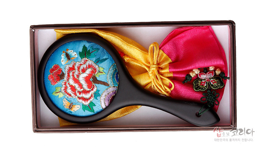 [텍스트: 샵오브코리아 대한민국의 품격까지 전합니다.] 하늘색 비단 바탕에 꽃과 나비 자수가 놓인 원형 손거울과 노랑·분홍색 비단 복주머니 세트 구성 이미지