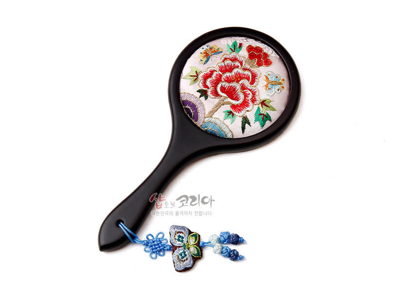 [텍스트: 샵오브코리아 대한민국의 품격까지 전합니다.] 연분홍 비단 바탕 위 붉은 모란과 나비 자수가 놓인 원형 손거울, 흑색 손잡이와 청색 나비 매듭 장식 구성