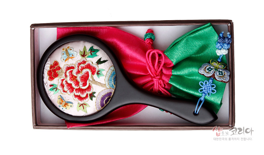[텍스트: 샵오브코리아 대한민국의 품격까지 전합니다.] 연분홍 비단 바탕 모란과 나비 자수 원형 거울, 흑색 나무 손잡이와 녹색·분홍 비단 주머니, 나비 매듭 장식 구성 이미지