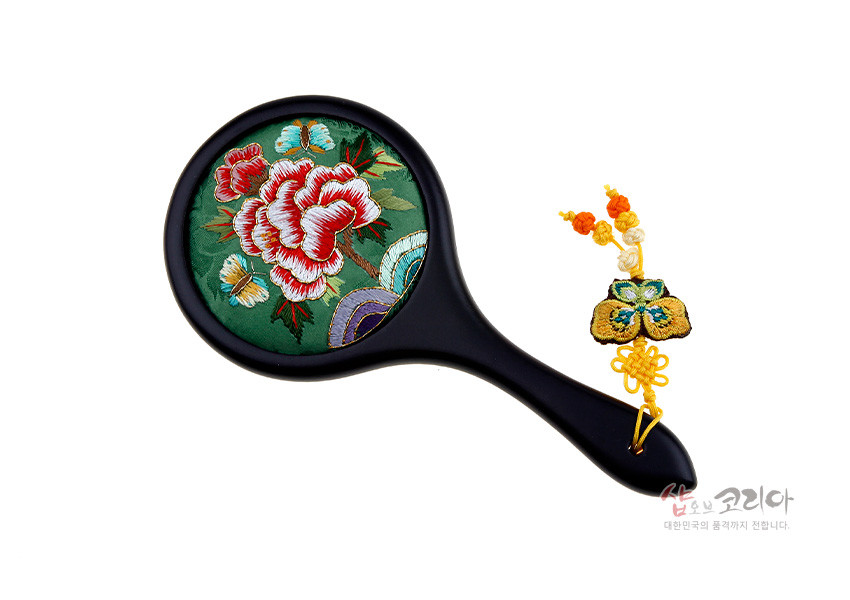 [텍스트: 샵오브코리아 대한민국의 품격까지 전합니다] 연두 비단 바탕에 붉은 모란꽃·나비 자수 장식 원형 손거울, 검은 손잡이와 노랑 매듭·복주머니 자수 노리개 장식 구성