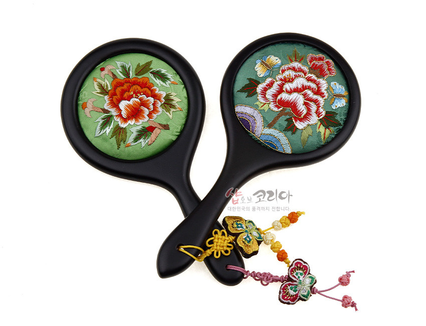 [텍스트: 샵오브코리아 대한민국의 품격까지 전합니다] 연두 비단 바탕 위 모란과 나비 자수 장식, 흑색 손잡이와 색실 노리개 매듭으로 포인트 준 전통 손거울 이미지 설명