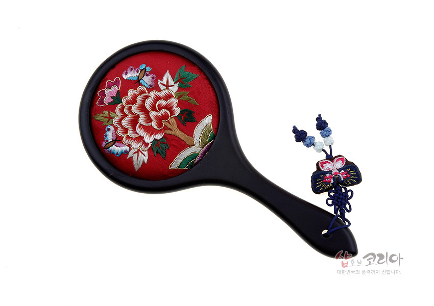 [텍스트: 샵오브코리아 대한민국의 품격까지 전합니다.] 붉은 비단 바탕에 모란과 나비 자수를 놓은 원형 손거울, 흑색 손잡이와 청색 매듭·복주머니 장식 술 구성