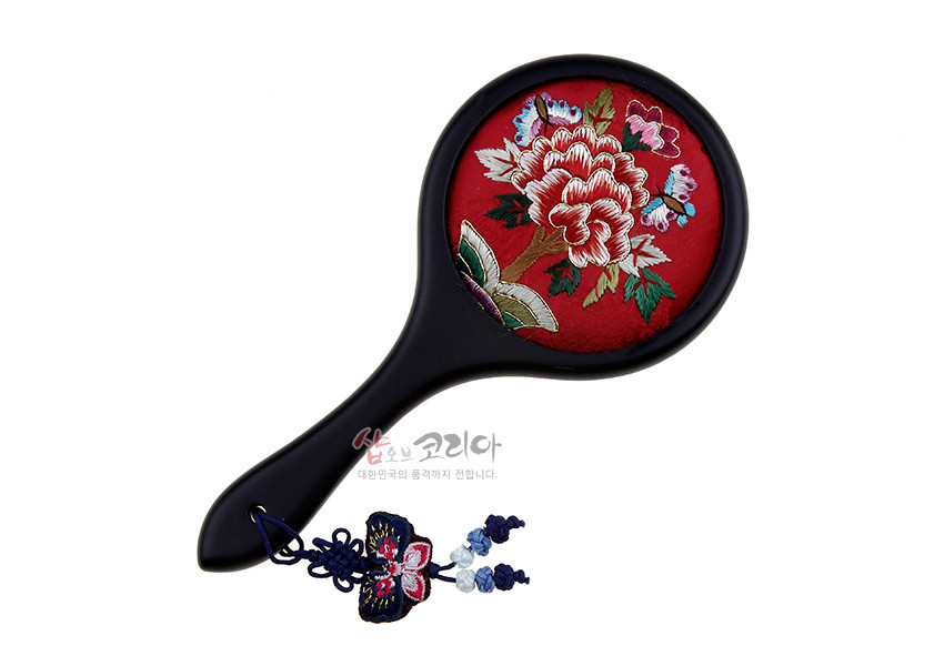 [텍스트: 샵오브코리아 대한민국의 품격까지 전합니다.] 붉은 비단 바탕 위 모란과 나비 자수 장식, 흑색 손잡이와 청색 매듭 노리개 술이 어우러진 손거울 이미지