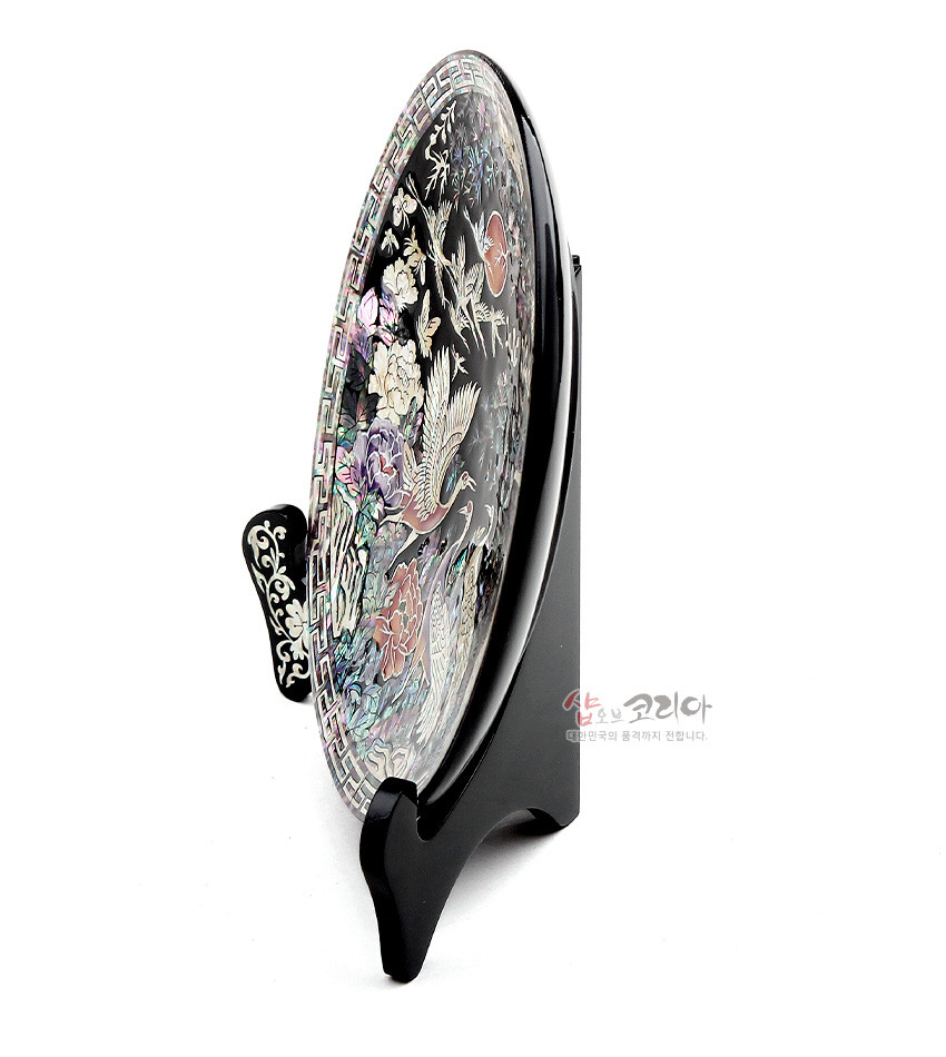 [텍스트: 샵오브코리아 대한민국의 품격까지 전합니다.] 흑색 옻칠 바탕 위 자개 상감으로 송학과 소나무, 꽃과 구름, 연속 기하학 문양을 정교하게 표현한 원형 쟁반과 받침대 장식품