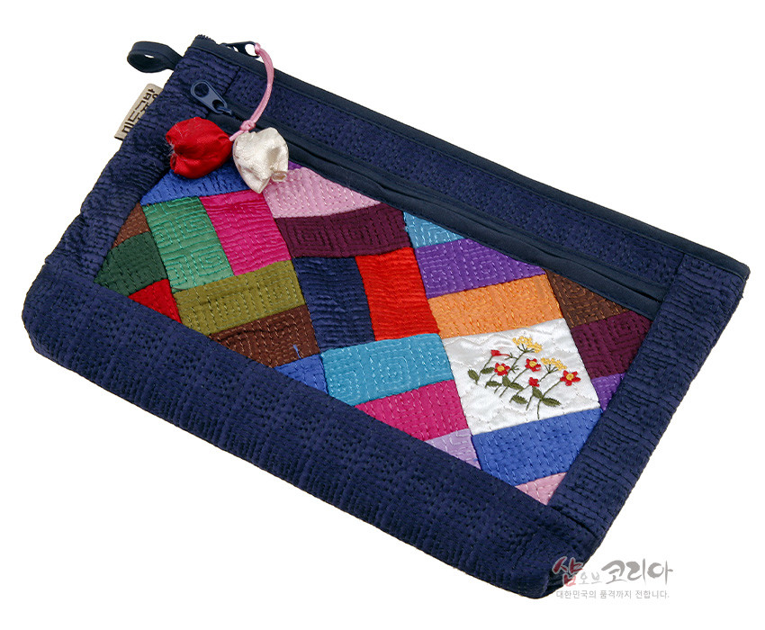 [텍스트: 샵오브코리아 대한민국의 품격까지 전합니다] 남색 누비 바탕에 다채로운 조각보 패치워크와 꽃자수 장식, 지퍼와 매듭 노리개 포인트가 어우러진 직사각 파우치 형태의 전통 누비 공예품