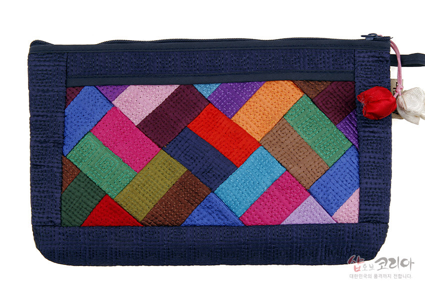 [텍스트: 샵오브코리아 대한민국의 품격까지 전합니다.] 남색 누비 바탕 위에 다채로운 조각보 누비 패턴이 더해지고 측면에 작은 매듭 장식이 달린 직사각형 파우치 형태의 디자인