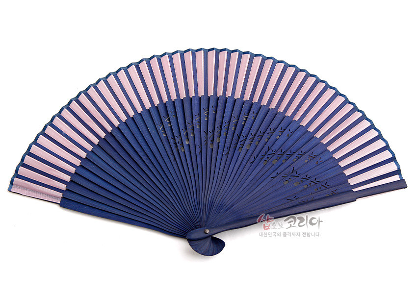 [텍스트: 샵오브코리아 대한민국의 품격까지 전합니다] 남색 나무 대와 분홍색 실크 접선이 조화를 이루고, 대 부분에 섬세한 식물 문양 투각 기법이 더해진 한국 전통 수공 부채 모습