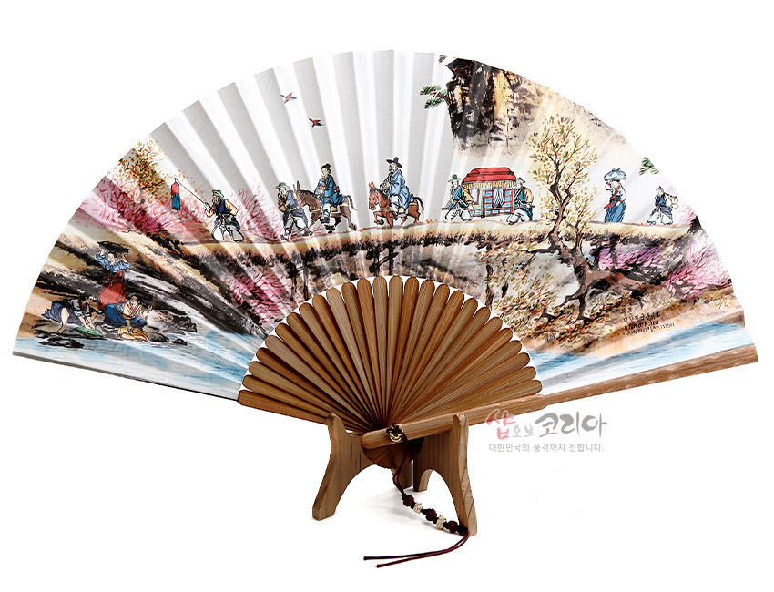 [텍스트: 샵오브코리아 대한민국의 품격까지 전합니다.] 대나무살과 흰 종이 위에 채색으로 표현된 전통 결혼 행렬과 산수 풍속도, 하단 물가 풍경과 나무 받침, 끈과 구슬 매듭 장식 부채 이미지 설명