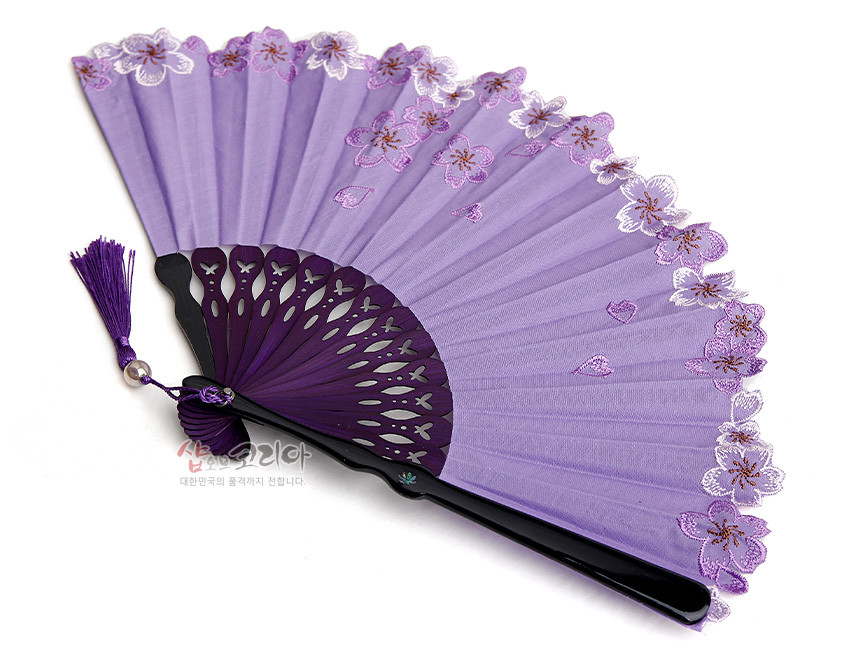 [텍스트: 샵오브코리아 대한민국의 품격까지 전합니다.] 연보라색 비단 선면에 자수 기법으로 수놓은 매화꽃과 레이스 가장자리 장식, 어두운 보라색 플라스틱 살대와 동색 장식 술이 조화를 이룬 전통 접이부채 모습