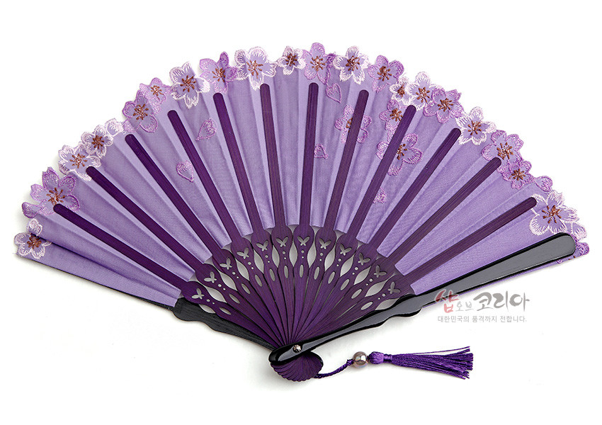 [텍스트: 샵오브코리아 대한민국의 품격까지 전합니다.] 연보라 비단 선면 위 자수 기법의 매화꽃 장식과 레이스 가장자리, 짙은 보라색 나무살과 술 장식이 조화를 이룬 전통 접이부채 이미지 설명
