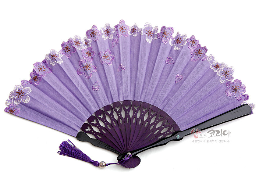 [텍스트: 샵오브코리아 대한민국의 품격까지 전합니다.] 연보라색 비단 선면에 보라색 매화 자수가 수놓인 레이스 가장자리와 투각 문양의 자주색 플라스틱 살대, 자색 술 장식 조합의 전통 접이부채 이미지 설명