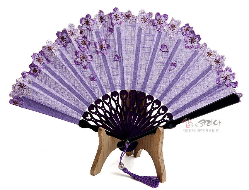 [텍스트: 샵오브코리아 대한민국의 품격까지 전합니다.] 연보라색 비단 선면에 자수 기법으로 수놓인 매화꽃과 레이스 가장자리, 자주색 플라스틱 살대와 장식 술이 어우러진 전통 접이부채 모습