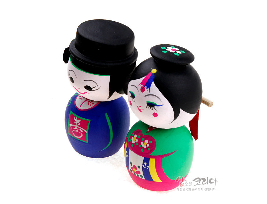 [텍스트: 喜, 샵오브코리아 대한민국의 품격까지 전합니다.] 원목 재질에 아크릴 채색 기법을 사용한 전통 혼례 신랑신부 목각 흔들인형, 원색 한복과 꽃문양 장식, 단아한 얼굴 표현의 디자인