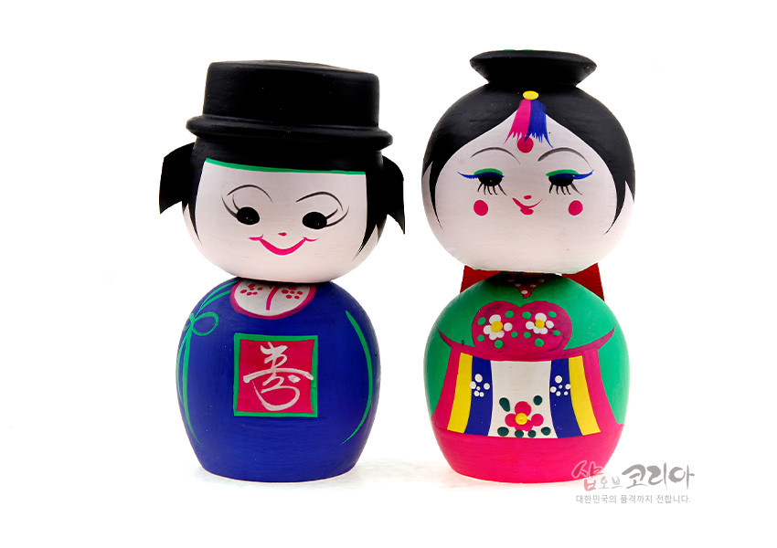 [텍스트: 喜, 샵오브코리아 대한민국의 품격까지 전합니다.] 원목 재질에 아크릴 채색 기법을 사용한 전통 혼례 신랑신부 목각 흔들인형, 원색 한복과 꽃·색동 문양이 돋보이는 디자인