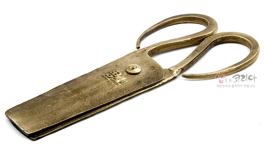 [텍스트: 복] 황동빛 금속 재질의 전통 엿가위, 두툼한 날과 곡선형 손잡이, 거친 표면 질감과 중앙의 한글 음각 문양이 돋보이는 공예품