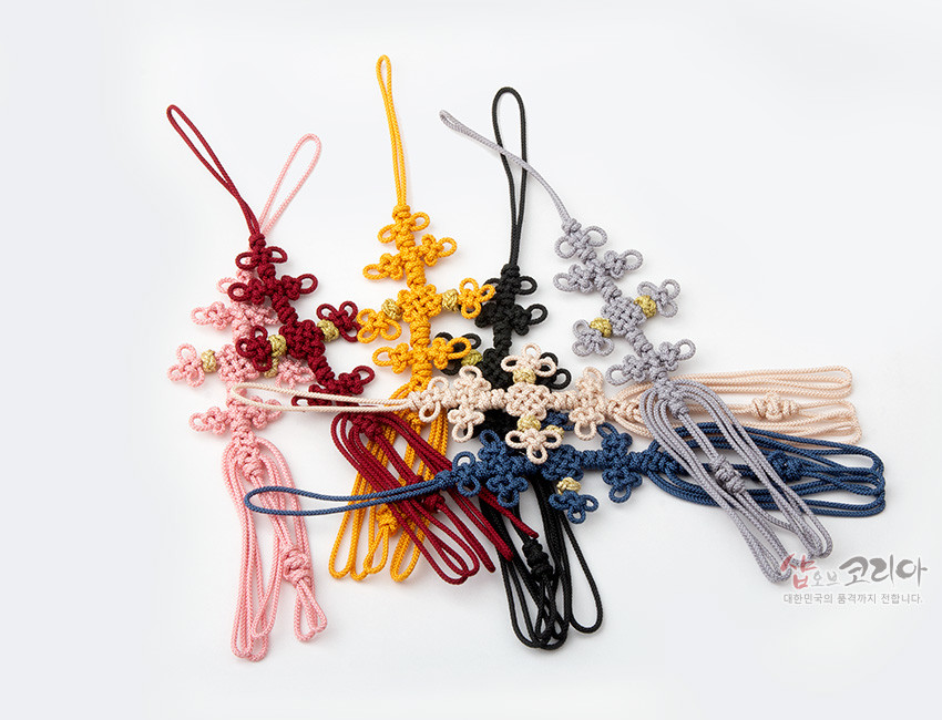 [텍스트: 샵오브코리아 대한민국의 품격까지 전합니다.] 합성실로 여러 겹 전통 매듭을 엮어 십자형 꽃문양을 이룬 작은 노리개, 다양한 색상의 실 조합과 길게 늘어진 술 장식 특징