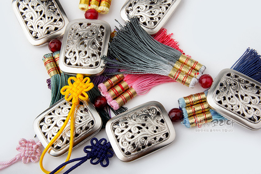 [텍스트: 샵오브코리아 대한민국의 혼을 세계까지 전합니다.] 은색 금속 사각 장식에 식물 투각 문양을 새기고, 다색 삼단 매듭 끈과 그러데이션 실숭어 술, 붉은 구슬 장식을 더한 작은 노리개 구성
