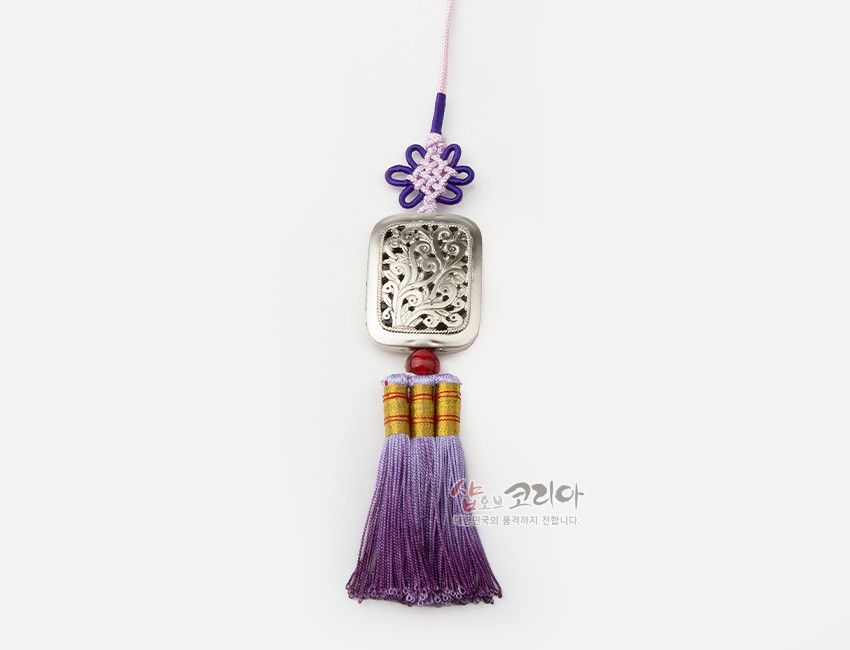 [텍스트: 샵오브코리아 대한민국의 혼, 존재까지 전합니다.] 은색 금속 사각 장식에 식물 문양 투각 기법, 자주색 전통 매듭과 보라 그러데이션 술로 구성된 작은 노리개 이미지 설명