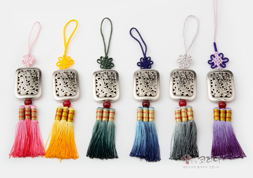 [텍스트: 샵오브코리아 대한민국의 품격까지 전합니다] 은색 금속 사각 장식에 식물 투각 문양과 붉은 구슬 장식, 다양한 색상의 전통 매듭과 그러데이션 술로 완성한 작은 노리개 구성