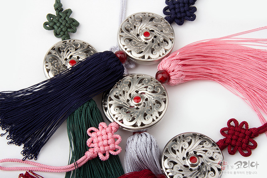 [텍스트: 샵오브코리아] 은색 금속 원형 당초무늬 장식 중앙 홍색 보석 장식, 남색·분홍·녹색 등 다색 전통 매듭과 실 술이 어우러진 작은 노리개 구성
