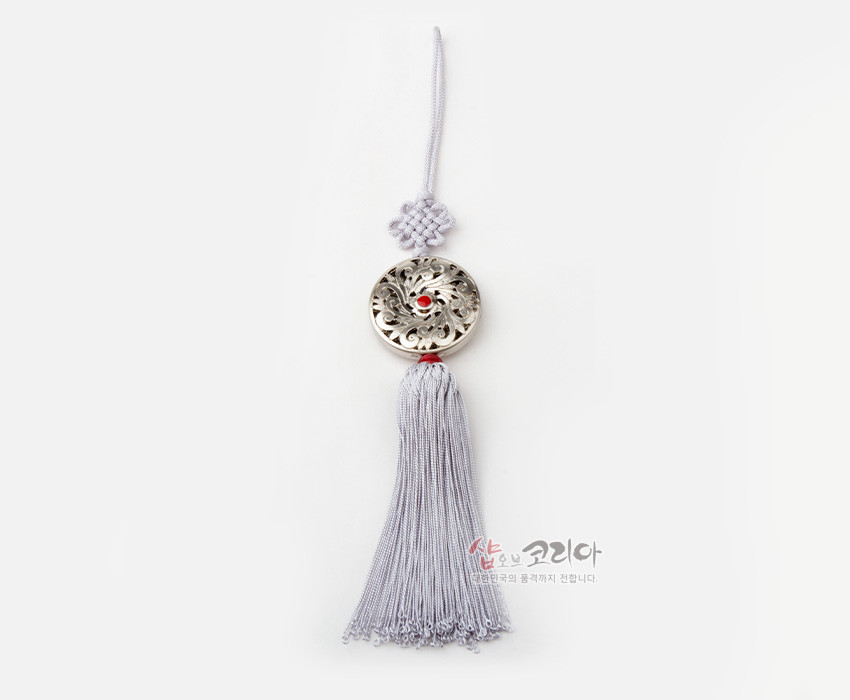 [텍스트: 샵오브코리아 대한민국의 공예가치 전합니다.] 은색 금속 원형 당초무늬 장식과 중앙 홍색 보석 장식, 연보라색 전통 매듭과 길게 늘어진 술로 구성된 작은 노리개 장식