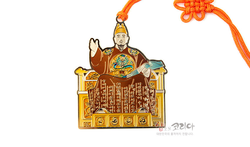 [텍스트: 샵오브코리아 대한민국의 품격까지 전합니다.] 금속 재질에 세종대왕과 한글 글귀를 채색한 장식, 하단 문양 장식과 주황색 전통 매듭 끈이 조화를 이룬 책갈피형 장식품