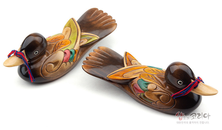 [텍스트: 샵오브코리아 대한민국의 품격까지 전합니다.] 채색 목각 기법으로 표현한 부부 원앙 한 쌍, 갈색 바탕에 노랑·초록·빨강 음각 문양과 빨강·파랑 매듭 장식 특징 이미지