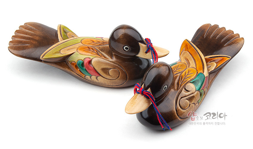 [텍스트: 샵오브코리아 대한민국의 품격까지 전합니다.] 원목 재질 부부원앙 목각 장식, 갈색 바탕에 녹색·주황·적색 채색 날개 문양과 전통 매듭 장식 특징 이미지