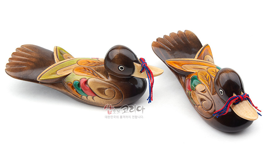 [텍스트: 샵오브코리아 대한민국의 품격까지 전합니다] 나뭇결이 살아있는 원목 재질, 갈색 바탕에 녹색·주황·적색 채색과 잎사귀 문양 조각, 부리에 빨강·파랑 전통 끈 장식 쌍원앙 목각 장식품 모습