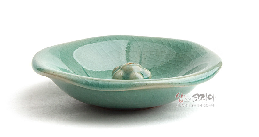 [텍스트: 샵오브코리아 대한민국의 품격까지 전합니다.] 비취색 청자 유약의 도자기 향꽂이, 연잎 음각 문양과 중앙 연밥 형상 장식이 조화를 이룬 연잎접시 디자인