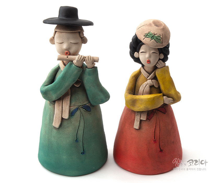 [텍스트: 샵오브코리아 대한민국의 혼과 정까지 전합니다.] 황토를 빚어 저온 채색한 도자기 인형 세트, 녹색 도포와 붉은 치마·노랑 저고리의 대비색과 단순화된 얼굴, 매무새를 살린 전통 한복 표현 인형 작품