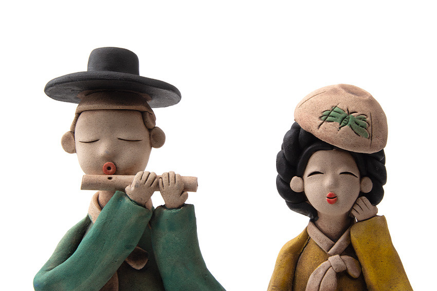 황토 재질로 구운 도자기 인형, 녹색 도포와 갓을 쓴 선비와 노랑 한복에 자수 모자 쓴 여인을 단순화된 얼굴과 은은한 채색으로 표현한 전통 공예품 이미지
