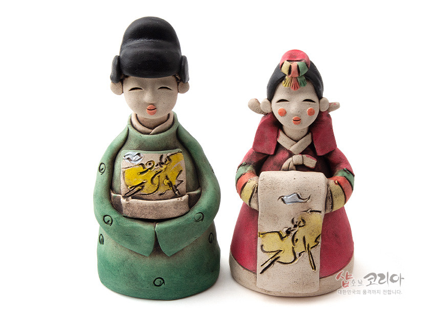 [텍스트: 샵오브코리아 대한민국의 품격까지 전합니다.] 황토를 빚어 저온 소성 후 채색한 도자기 신랑신부 인형, 녹색·홍색 한복과 전면 노란 바탕 학·구름 추상 문양이 조화된 전통 혼례 조형물