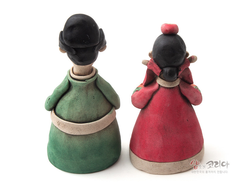 [텍스트: 샵오브코리아 대한민국 전통의 품격까지 전합니다.] 황토 도자 재질로 빚은 전통 혼례 신랑·신부 인형의 후면, 녹색·붉은색 한복 채색과 단정한 머리 모양이 돋보이는 조형미 표현