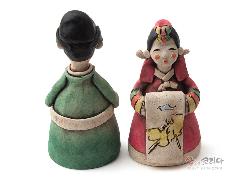 [텍스트: 샵오브코리아 대한민국의 품격까지 전합니다.] 황토 재질 도자 기법의 신랑신부 인형, 녹색·홍색 한복과 수묵화풍 노란 동물 문양 두루마리 장식이 어우러진 혼례 조형품 모습