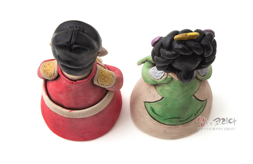 [텍스트: 샵오브코리아 대한민국의 품격까지 전합니다.] 붉은 곤룡포와 녹색 활옷을 입은 황토 도자기 왕과 왕비 인형 후면, 입체 머리장식과 채색 한복 장식이 돋보이는 구도