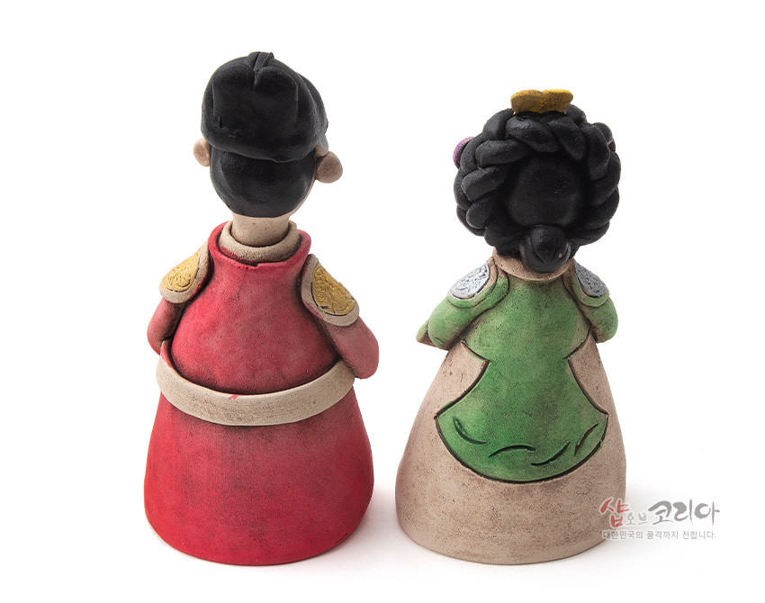 [텍스트: 샵오브코리아 대한민국의 품격까지 전합니다.] 붉은 곤룡포와 녹색 대례복을 입은 황토 도자 왕과 왕비 인형의 후면, 양각 견장과 묵직한 머리 비녀 장식이 돋보이는 전통 조형물 표현