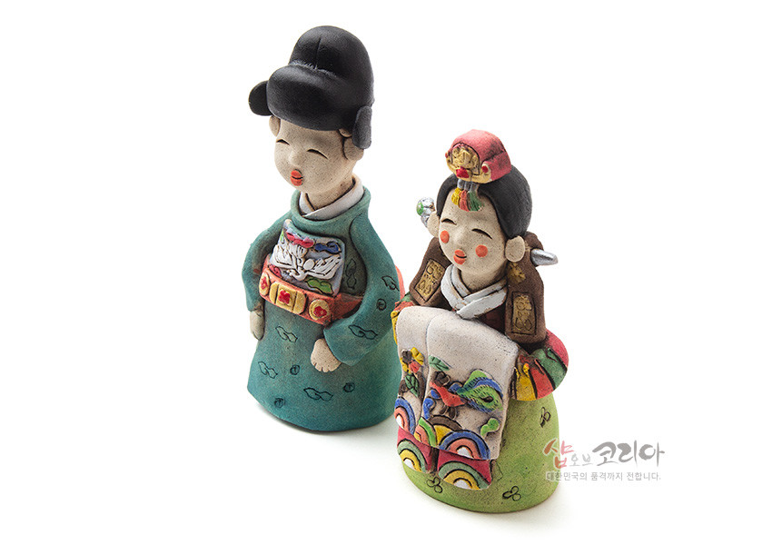 [텍스트: 샵오브코리아 대한민국의 혼까지 전합니다.] 채색 황토 도자기 재질로 빚은 전통 혼례복 신랑신부 인형, 오방색 동정과 활옷, 수복·봉황·무지개 문양을 손채색으로 표현한 공예품