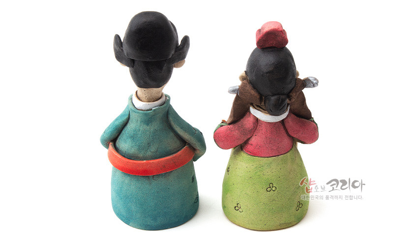 [텍스트: 샵오브코리아 대한민국의 품격까지 전합니다.] 채색 황토 재질의 신랑신부 도자기 인형, 청록·홍·연두색 대비와 단정한 머리 장식, 삼엽 문양이 있는 뒷모습 표현 작품