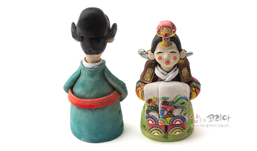 [텍스트: 샵오브코리아 대한민국의 품격까지 전합니다.] 채색 황토 재질의 도자기 기법 신랑신부 인형, 오방색 혼례복과 봉황·무지개·길상무늬를 입체적으로 표현한 전통 공예품 묘사