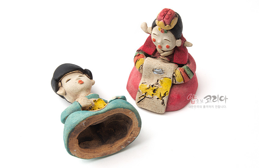 [텍스트: 샵오브코리아 대한민국의 품격까지 전합니다.] 황토를 성형·소성한 도자기 인형, 붉은색과 청록색 채색의 전통 혼례복과 추상화된 노란 수묵화풍 그림 장식이 어우러진 신랑신부 한쌍 인형 구성